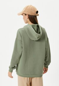 Sudadera holgada de color verde oliva con capucha ajustable, puños y dobladillo acanalados. Combinada con una gorra de béisbol de piel sintética color beige. Diseño simple.