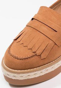 Mocassin en daim marron avec détail à franges sur le dessus et bordure en corde tissée autour de la semelle compensée.