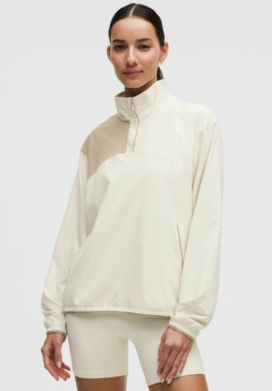 Frau trägt einen hochgeschlossenen Pullover in Beige und Creme mit langen Ärmeln und passende, enganliegende Shorts vor schlichtem Hintergrund.