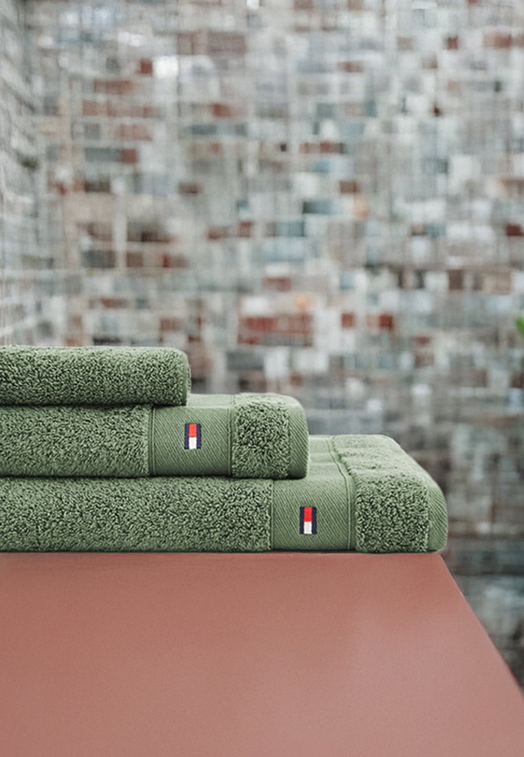 Zielone ręczniki w różnych rozmiarach, starannie ułożone, z teksturowanej tkaniny i prostokątnym kształcie; każdy z małym, czerwono-białym-niebieskim logo.