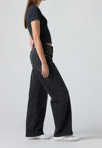Personne portant un t-shirt court noir, un jean noir ample et des baskets blanches, debout de côté devant un fond gris.