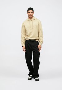 Felpa beige con cappuccio e lacci, abbinata a pantaloni cargo neri con tasche laterali. Calzature comprendono scarpe nere con dettagli bianchi.