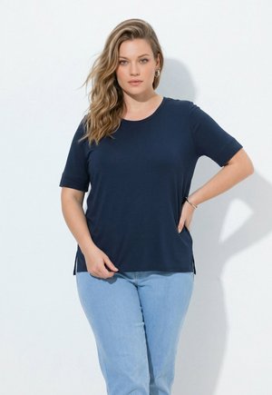 Frau mit welligem blondem Haar, die ein dunkelblaues kurzärmeliges Shirt und hellblaue Jeans trägt, steht mit einer Hand in der Hüfte.