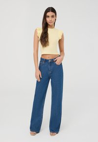 Top corto giallo chiaro con maniche a volant; jeans a vita alta blu a gamba larga; modello che indossa sandali con tacco beige.