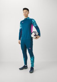 Puma Sportanzug in Türkis mit pinken Akzenten, besteht aus einem Reißverschlussoberteil und einer figurbetonten Hose mit geometrischen Mustern; das Model hält einen Fußball.