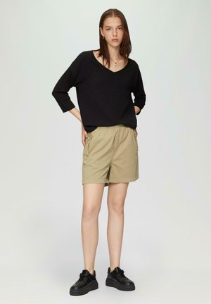 Schwarzer V-Ausschnitt-Pullover, khakifarbene Shorts mit seitlichen Taschen, umgeschlagenem Saum und schwarzen Turnschuhen. Minimalistisches Design und lässige Passform.