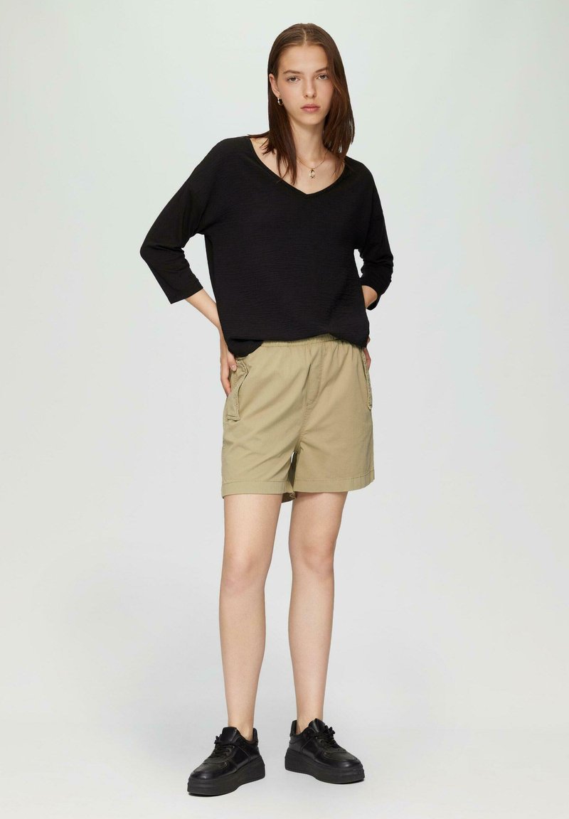 Schwarzer V-Ausschnitt-Pullover, khakifarbene Shorts mit seitlichen Taschen, umgeschlagenem Saum und schwarzen Turnschuhen. Minimalistisches Design und lässige Passform.