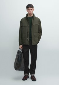 Giacca verde oliva con due tasche sul petto, indossata sopra un maglione verde scuro, abbinata a pantaloni neri e scarpe marroni, con in mano una borsa grigia.