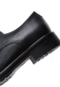 Zapato de cuero negro con textura suave, forma redondeada y suela de goma resistente con un patrón de tracción. Diseño minimalista, sin adornos.