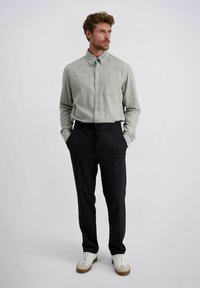 Man met handen in zijn zakken, gekleed in een lichtgrijze overhemd met knopen, zwarte drawstring broek en witte sneakers met beige zolen, staande tegen een witte achtergrond.