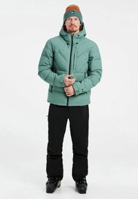 Chaqueta acolchada verde con capucha, cremallera negra, bolsillos laterales y un acabado texturizado; combinada con pantalones negros y botas de esquí negras.