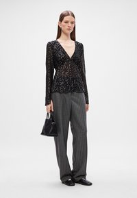 Μαύρο διαφανές τοπ με μακριά μανίκια, λουλουδάτη κεντημένη διακόσμηση και βαθύ V-λαιμό, συνδυασμένο με γκρίζες ριγέ παντελόνες φαρδιάς εφαρμογής και μια μικρή μαύρη τσάντα.