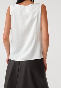 Mujer con una blusa blanca sin mangas y una falda negra, vista desde atrás sobre un fondo liso.