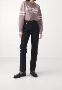 Maglione corto viola con testo bianco "Coach New York" e strisce. Jeans neri a vita alta. Mocassini neri con finitura testurizzata.