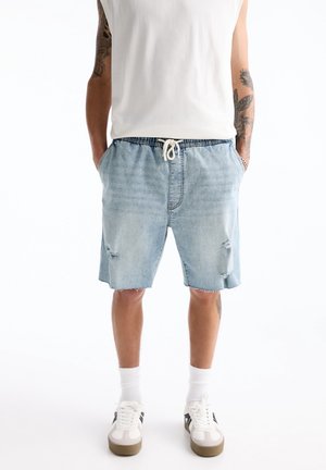 Homme portant un short en denim bleu clair avec un cordon, des chaussettes blanches, des baskets blanches et un débardeur blanc, debout les mains dans les poches.