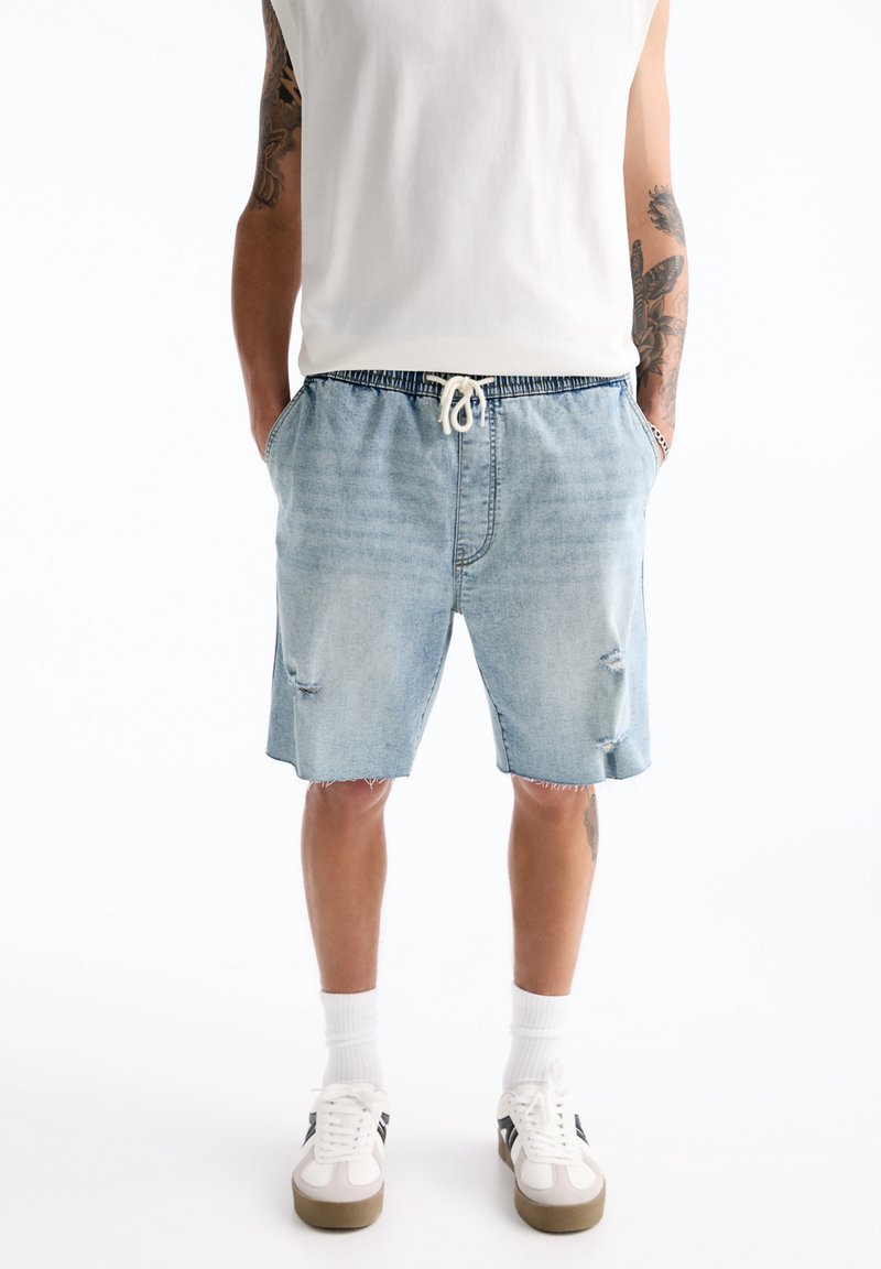 Homme portant un short en denim bleu clair avec un cordon, des chaussettes blanches, des baskets blanches et un débardeur blanc, debout les mains dans les poches.