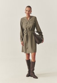 Olivengrünes, langärmliges Kleid mit V-Ausschnitt, zwei Brusttaschen und einem gebundenen Taillengürtel. Kombiniert mit dunklen kniehohen Stiefeln und einer strukturierten Handtasche.