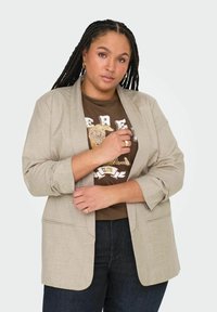 Helehall blazer pehmealt struktureeritud disainiga, rullitud varrukad, küljetaskud, kantud tumeda pruuni graafilise T-särgi ja tumedate teksade peal.