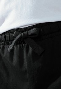 Black cotton joggers feature an elastic waistband with a textured drawstring. A white shirt is partially visible above the waistband.