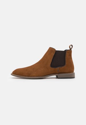 Stivale Chelsea in suede marrone con pannelli elastici neri e suola in pelle, mostrato dal lato esterno su sfondo bianco.