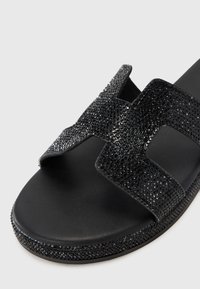 Svarta slide-sandaler med texturerade remmar prydda med små, glänsande svarta kristaller. Texturerad fotbädd och slät sula.