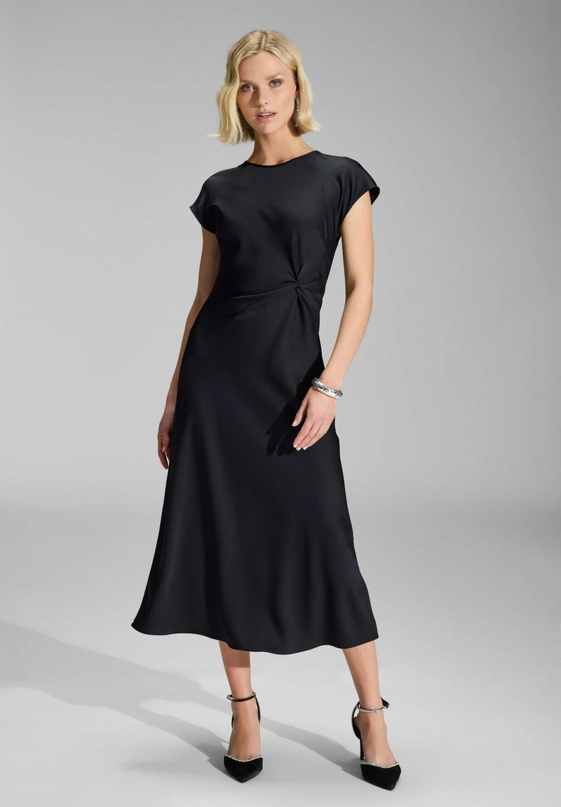 Sort satin midi-kjole med knudedetalje i taljen, korte ærmer og en glat, flagrende nederdel. Sammen med sorte stilletter og et sølvarmbånd.