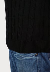 Fekete kötött pulóver texturált kábelmintával, bordás aljjal és mandzsettákkal, világoskék farmernadrággal párosítva.