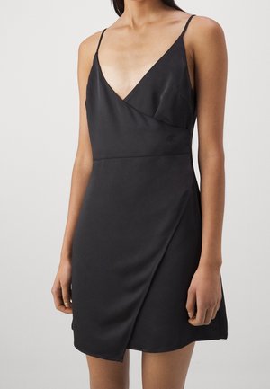 Femme portant une robe portefeuille noire sans manches avec fines bretelles et un décolleté en V, debout devant un fond clair uni.