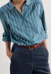 Blue striped button-up shirt tucked into navy corduroy pants with a brown braided leather belt and hands in pockets.