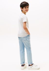 Camiseta gris de manga corta, jeans de mezclilla azul claro y zapatillas blancas con acentos en marino y rojo, niño de pie con las manos en los bolsillos.