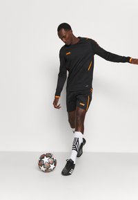 Completo sportivo nero con accenti arancioni, composto da una maglia a maniche lunghe e pantaloni corti, abbinato a scarpe da calcio nere e un pallone da calcio.