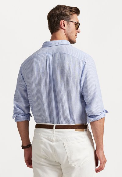 Gaiši zilas svītrainas krekls ar pogām un sarullētām piedurknēm, izgatavots no elpojoša auduma, kombinēts ar baltām biksēm un brūnu jostu.