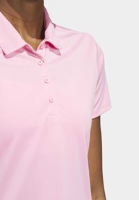 Gros plan sur une personne portant un polo à manches courtes rose clair avec col et trois boutons, sur un fond gris clair uni.