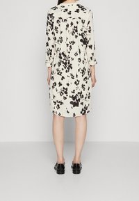 Kreem ja musta lillemustriga kleit, millel on kolmveerandvarrukad, lõdva istuvuse ning volditud detail taga. Seljas on mustad jalanõud.