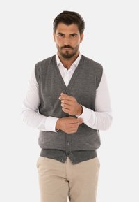 Gilet grigio lavorato a maglia con scollatura a V, chiusura con bottoni e orlo a coste. Indossato sopra una camicia bianca, con pantaloni beige. Design semplice e solido.