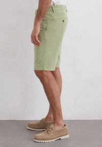 Light green shorts with pockets and a back button detail, paired with tan suede boat shoes and textured soles. Side view, standing posture.