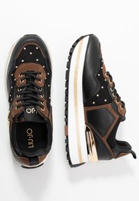 Svarta och bruna sneakers med en strukturerad ovandel, guldiga accenter och en tjock vit och guld sula, som har dekorativa nitar och en siddragkedja.