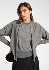 Cardigan gris de punto con frente abierto, mangas largas y puños acanalados. Combinado con una camiseta gris de cuello redondo y una falda negra. Textura suave, diseño minimalista.
