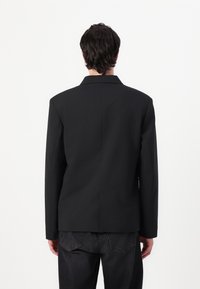 Schwarzer Blazer mit strukturiertem Design, der einen Reverskragen, lange Ärmel und eine klare Silhouette aufweist; der Stoff erscheint glatt und tailliert.