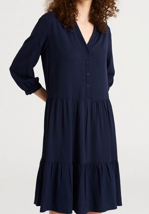 Mujer con un vestido azul marino, de manga larga, con volantes, escote en V y cierre de botones en la parte delantera.