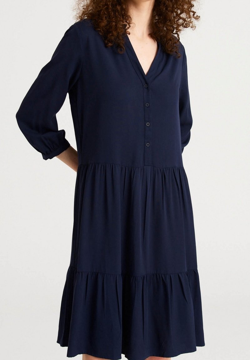 Femme portant une robe bleu marine à manches longues, à volants, avec un décolleté en V et une patte de boutonnage à l'avant.