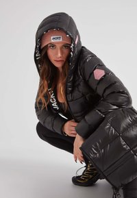 Mujer agachada con una chaqueta de plumas negra con capucha, gorro rosa etiquetado "JACK IT", leggings negros y botas de senderismo negras con detalles amarillos.