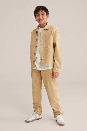 Chaqueta de pana beige con un bolsillo de parche, combinada con pantalones a juego. Ambos tienen una textura acanalada. Camiseta blanca debajo y zapatillas blancas.