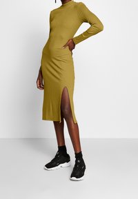 Olivgrünes geripptes Strickkleid mit hohem Kragen und seitlichen Schlitzen; kombiniert mit schwarzen Sportschuhen und Socken. Glatte Textur und figurbetontes Design.