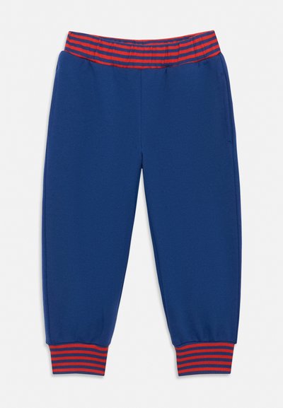 Modré detské tepláky s elastickým pásom a manžetami, ozdobené červenými a modrými horizontálnymi pruhmi na bielom pozadí.