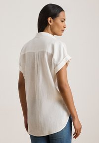 White linen shirt with short rolled sleeves, a back yoke seam, and a relaxed fit. Worn with blue jeans; textured fabric evident.