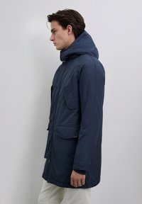 Ung mand i en navyblå hættetrøje og lyse bukser, som står sidelæns mod en ensfarvet lys grå baggrund.