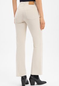 Vrouw in beige corduroy rechte pijpenbroek en zwarte leren enkellaarsjes, gezien vanachter tegen een witte achtergrond.