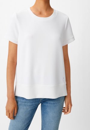 Biała bluzka z krótkim rękawem o okrągłym dekolcie, charakteryzująca się lekko oversizedowym krojem i gładką fakturą, zestawiona z dżinsami.