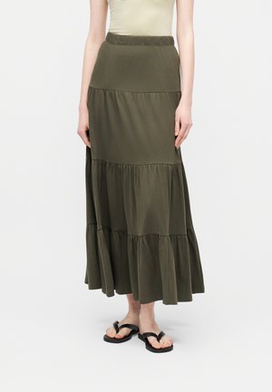 Femme portant une jupe longue à volants couleur olive, un haut beige sans manches, et des sandales à anneau pour orteils noires, debout sur un fond blanc uni.